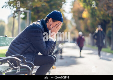 Giovane uomo preoccupato è che copre il suo volto. Egli è seduta su una panchina durante la soleggiata giornata autunnale. Donna e una ragazza visibile in background. Foto Stock