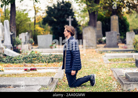 Lutto giovane uomo in ginocchio di fronte a una grave su un cimitero durante un triste giorno d'autunno. Foto Stock