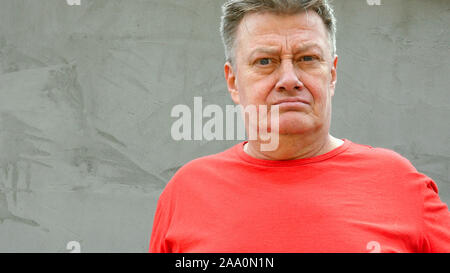 Ritratto di grave senior uomo adulto in t-shirt rossa, chi sta guardando la telecamera. Contro lo sfondo grigio del muro intonacato in stile loft design. Foto Stock