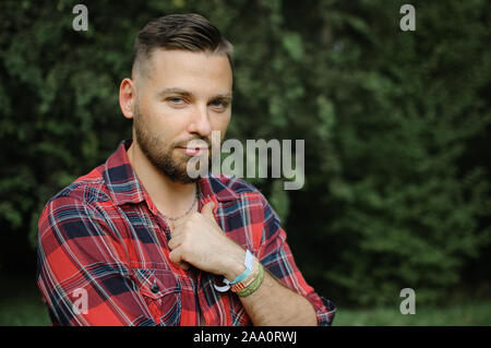 Ritratto all'aperto di rilassata bel giovane uomo con la barba nel parco su alberi verde dello sfondo. Tanga concetto di stile. Foto Stock