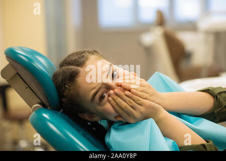 Bambina nella paura chiude la sua bocca con le sue mani seduto in una sedia nello studio dentistico. Foto Stock
