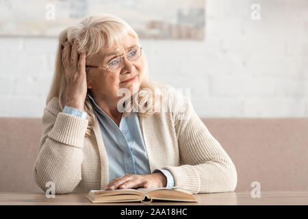 Sconvolto senior donna in bicchieri seduto vicino a libro Foto Stock