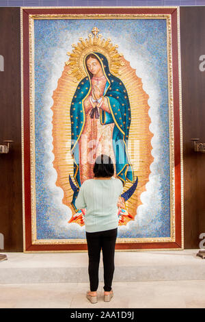 Un ispanico adoratore ammira un ritratto della Madonna di Guadalupe per artista Valerio Lenarduzzi in una cattedrale in arancione, CA. La Vergine di Guadalupe (Spagnolo: Virgen de Guadalupe), è un cattolico a titolo della beata Vergine Maria associato ad una apparizione mariana. Foto Stock