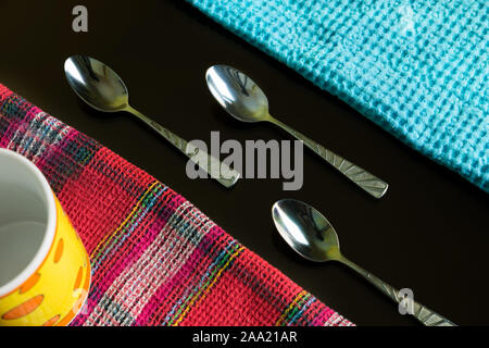 Tre cucchiaini in argento tra tovaglioli blu e multicolore con tazza gialla sullo sfondo scuro riflettente. Linee e posate blu e colorate. Foto Stock