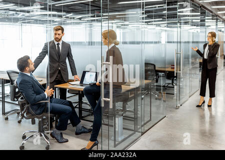 Gruppo di rigorosamente vestito la gente di affari avente una discussione, seduto sul posto di lavoro con un computer in un ufficio moderno con pareti divisorie in vetro Foto Stock