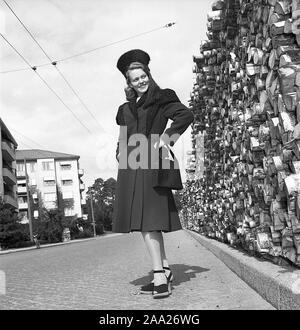 Le donne della moda nel 1940s. Una giovane donna in un tipico degli anni quaranta rivestire con abbinamento di hat, le scarpe e la borsetta. È il dopoguerra in Svezia e non vi è ancora una mancanza di combustibile e carbone per il riscaldamento e le cataste di legna da ardere è visibile in background. Kristoffersson Ref V78-6. La Svezia 1947 Foto Stock