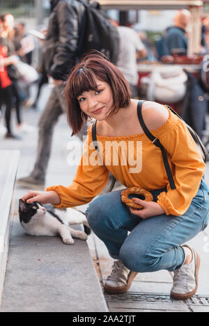 Giovane e bella ragazza in abiti alla moda tocca e ama un homeless street gatto a una strada trafficata. Foto Stock