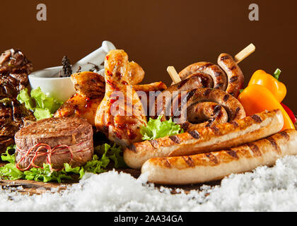 Piccante deliziose grigliate di carne per un inverno di barbecue con una chiusura di un medaglione di filetto di maiale e salsicce di manzo e le cosce di pollo su un letto di fresco Foto Stock