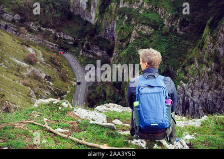 Vista posteriore giovane escursionista con zaino seduto sulla vetta della montagna godendo il paesaggio di rocce e auto sfondo su strada. Lo stile di vita di viaggio wanderlust avvento Foto Stock