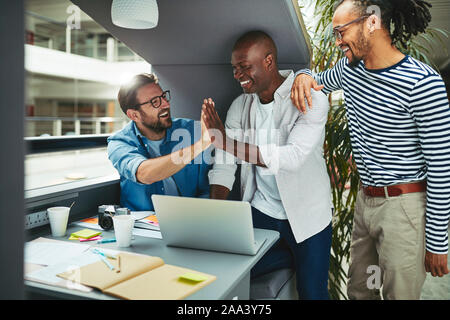 Ridendo gruppo di diversi professionisti creativi di alta fiving ogni altro mentre si lavora su un computer portatile insieme in un ufficio incontro pod Foto Stock