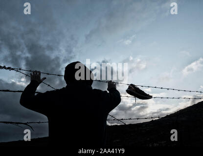 Vista posteriore dell'uomo felpa con cappuccio da indossare dietro il filo spinato, filo di rasoio recinzione. Calzatura appesi da recinzione Foto Stock