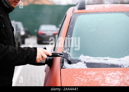 L'uomo cancella la neve ghiacciata dal parabrezza della vettura. Il raschiatore in Le Mans mano. Pulizia finestrino congelate di orange automobile. Foto Stock