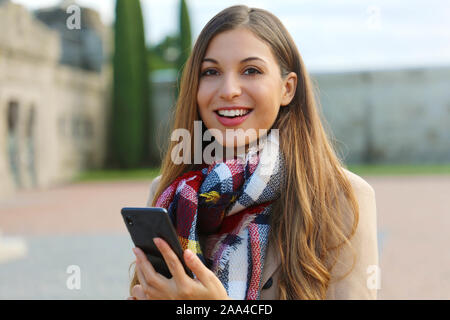 Felice allegro giovane donna guarda a voi in possesso di telefono cellulare sulla piazza della città indossando cappotto e sciarpa in inverno. Foto Stock