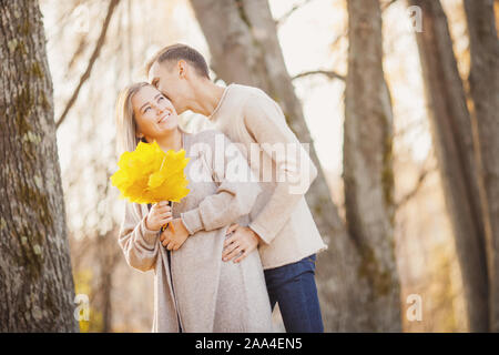 Amare giovane in autunno park con foglie di acero, uomo sussurra a bella ragazza orecchio dichiarazione d'amore. Foto Stock