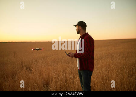 Agricoltore detiene remote controller con le sue mani mentre quadcopter è volare su sfondo. Drone vola dietro l'agronomo nel campo di grano Foto Stock