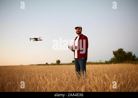 Agricoltore detiene remote controller con le sue mani mentre quadcopter è volare su sfondo. Drone vola dietro l'agronomo nel campo di grano Foto Stock