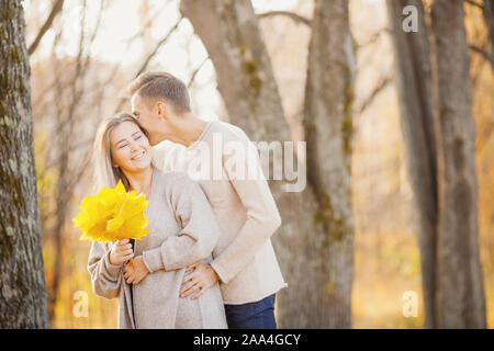 Amare giovane in autunno park con foglie di acero, uomo sussurra a bella ragazza orecchio dichiarazione d'amore. Foto Stock