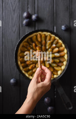 Donna slanciata mano con miele bilanciere sopra in casa a forma di fiore torta di prugne in ghisa pan su sfondo nero in vista dall'alto uno spazio di copia Foto Stock