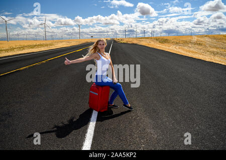 Attraente donna bionda con bagagli rosso autostop su un vuoto che la strada di campagna Foto Stock