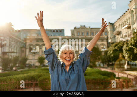 Età non costituisce una barriera. Ritratto di felice e bella donna senior sollevando in alto le mani e sorridenti mentre in piedi all'aperto in una giornata di sole. La vecchiaia. Felicità.. Foto Stock