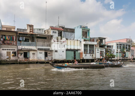 In Cai Be, il Delta del Mekong, Vietnam - Marzo 13, 2019: lungo quello dei Kinh 28 canal. 2 ragazzi sul basso battello motorizzato su acqua marrone passando nella parte anteriore della fila di abitazioni Foto Stock