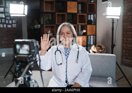 Saluti femmina senior medico con un alto cinque per registrare un video del suo blog Foto Stock