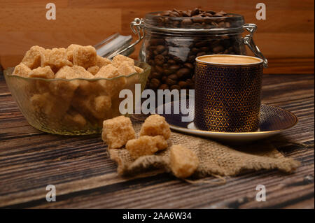 Una tazza di caffè, i chicchi di caffè in un vasetto di vetro e pezzi di zucchero di canna su uno sfondo di legno Foto Stock