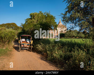 Un tradizionale oxcart di Bagan, Myanmar rotola giù una strada sterrata nella zona archeologica con stupa e un grande tempio a distanza Foto Stock