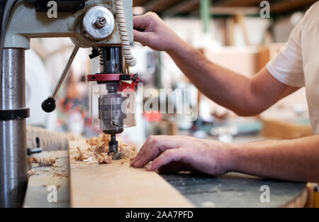 Carpenter lavorando su una potenza verticale praticare la perforazione di un foro in un asse di legno in un vicino sul bit, segatura e le sue mani Foto Stock