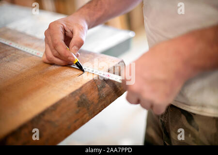 Falegname tenendo la misurazione con un nastro di misurazione la marcatura della lunghezza su un asse di legno con un marcatore in un vicino fino alle mani Foto Stock