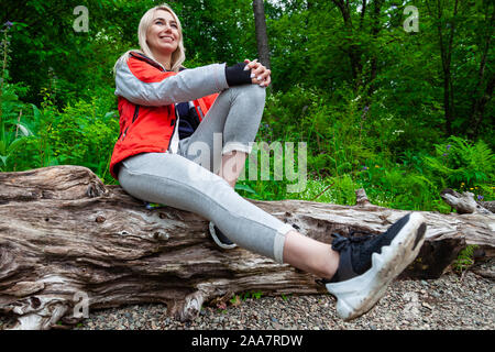 Bella ragazza bionda in una camicia d'autunno con un zaino rosso si siede su un albero caduto in un insieme di strutture o in un parco in natura, passeggiate e viaggiano in aria fresca. Foto Stock