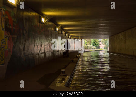 Figura isolata e solitaria che cammina il cane dal canale urbano nel sottopassaggio pedonale britannico scuro, passando opere d'arte di graffiti urbani spruzzate sulle pareti. Foto Stock