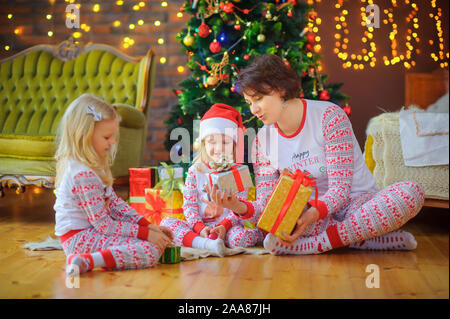 Mom fornisce doni ai bambini seduti sul pavimento vicino la festosa albero di Natale. La mattina di Natale Foto Stock