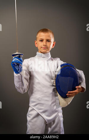 Little Boy in un bianco tuta protettiva maschera da scherma e con esercizi di pinza nella scherma su sfondo scuro Foto Stock