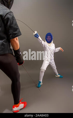 Giovane ragazza bionda in un coaching scuro vestito e un ragazzino in un bianco vestito sportivi praticanti di scherma di pinza su sfondo grigio Foto Stock