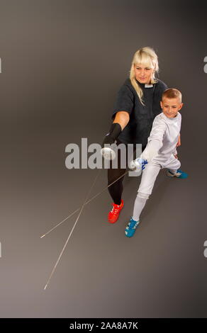 Giovane ragazza bionda in un coaching scuro vestito e un ragazzino in un bianco vestito sportivi praticanti di scherma di pinza su sfondo grigio Foto Stock