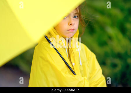 Carino bambina in un impermeabile giallo è nascosto dalla pioggia sotto un ombrello Foto Stock