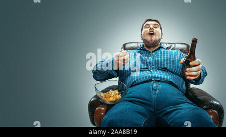 Ritratto di grasso uomo caucasico indossando jeanse e whirt seduto in una poltrona marrone isolato sul gradiente dello sfondo grigio. Guardando la TV bevande birra, mangia patatine e cambiare i canali. Il sovrappeso e spensierato. Foto Stock