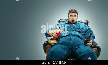 Ritratto di grasso uomo caucasico indossando jeanse e whirt seduto in una poltrona marrone sul gradiente dello sfondo grigio. Guardando la TV bevande birra, mangia patatine, patate fritte. Il sovrappeso e spensierato. Foto Stock