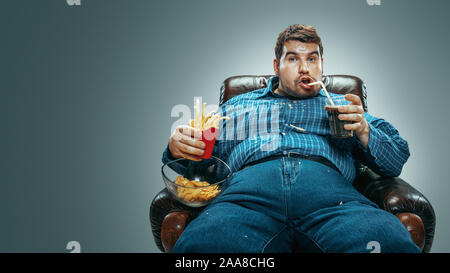 Ritratto di grasso uomo caucasico indossando jeanse e whirt seduto in una poltrona marrone sul gradiente dello sfondo grigio. Guardando la TV le bevande cola, mangia patatine, patate fritte, laughting. Il sovrappeso e spensierato. Foto Stock