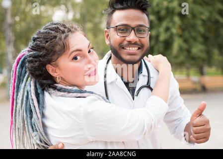 Indian giovane medico con occhiali, bello. Abbraccia una donna bianca medico. Giovani uomini in camici avvolgente, per esterno Foto Stock