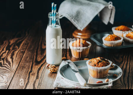 Vaniglia e caramello muffin in bicchieri di carta e bottiglie di latte sul legno scuro dello sfondo. Deliziosa tortina con uvetta, mandorle e noci. In casa biscui Foto Stock