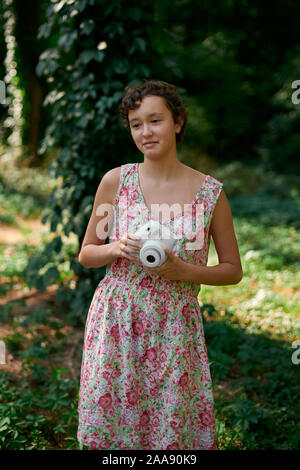 Adorabile ragazza preteen tenendo la fotocamera istantanea e prendendo foto chiudendo gli occhi nel parco verde Foto Stock