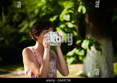 Entusiasta ragazza preteen sollevamento fotocamera istantanea e prendendo la foto in parco verde Foto Stock