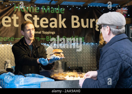 Market Trader serve un cliente con un Burger di bistecca con pudding nero e formaggio in un panino al York Christmas Market, North Yorkshire, Regno Unito. Foto Stock