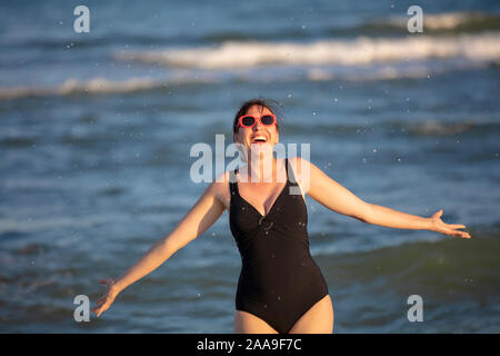 Donna felice al mare. Donna in costume da bagno schizza acqua su uno sfondo di onde. La vacanza estiva sulla spiaggia. Foto Stock