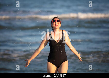 Donna felice al mare. Donna in costume da bagno schizza acqua su uno sfondo di onde. La vacanza estiva sulla spiaggia. Foto Stock