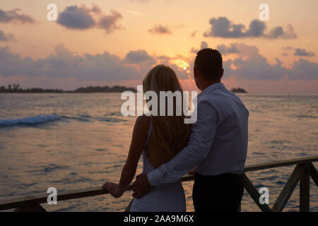 Vista posteriore di appena sposato abbracciarsi e guardare il tramonto skyline sull isola tropicale sulla spiaggia di Maldive. Oceano turchese laguna sullo sfondo Foto Stock