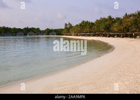 Costa dell'oceano indiano sulle isole Maldive. Sedie a sdraio sotto gli ombrelloni di paglia. Spiaggia di sabbia bianca e mare calmo. Bungalows in background Foto Stock