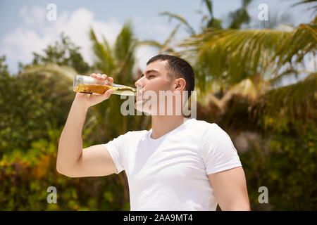 Giovane uomo beve birra sulla spiaggia tropicale con palme di cocco sullo sfondo. Maschio caucasico con una bottiglia di limonata bevanda disseta la sete Foto Stock